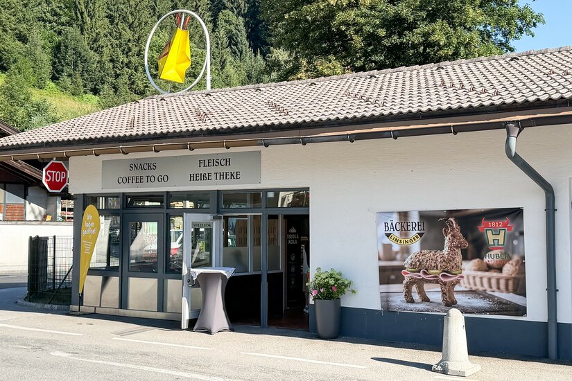 Außenansicht Imbissfiliale Linsinger Außenansicht einer Bäckerei- und Imbissfiliale mit Werbeplakaten und Stehtisch vor dem Eingang.