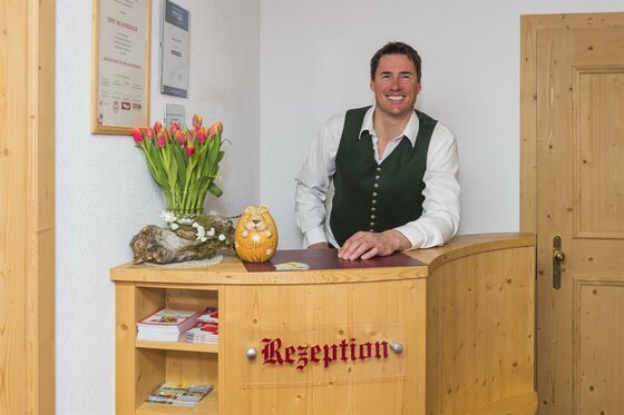 Ein Mann in Trachtenweste steht hinter einem hölzernen Empfangstresen mit der Aufschrift Rezeption.