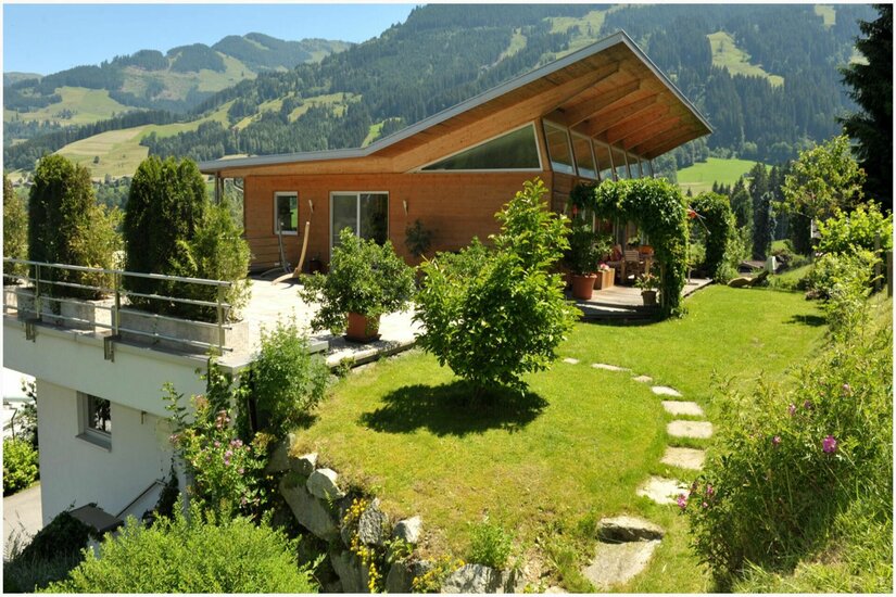 Energetische Praxis im Grünen Modernes Holzhaus mit Pultdach und Garten vor bewaldeten Bergen.