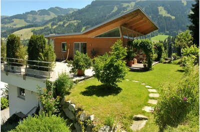 Energetische Praxis im Grünen Modernes Holzhaus mit Pultdach und Garten vor bewaldeten Bergen.
