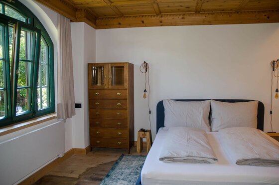 Schlafzimmer mit Doppelbett, Holzkommode und Rundbogenfenster unter einer Holzdecke.