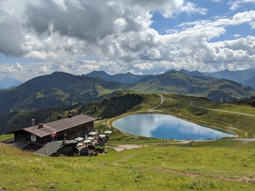 Berghütte am Speichersee Berghütte mit Terrasse neben einem runden Speichersee in einer alpinen Graslandschaft vor Gebirgskette.
