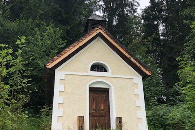 Plattenkapelle Eine kleine Kapelle inmitten von dichten Bäumen. Das Gebäude hat eine hellbeige Fassade und ein steiles Dach.
