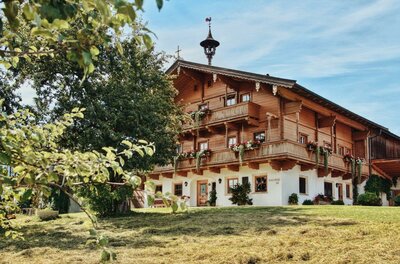 Ein traditionelles, chaletartiges Haus mit mehreren Balkonen und Blumenschmuck. Umgeben von Grünflächen und Bäumen unter einem klaren Himmel.