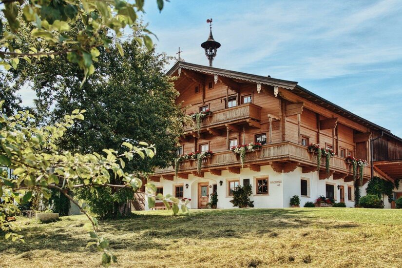 Ein traditionelles, chaletartiges Haus mit mehreren Balkonen und Blumenschmuck. Umgeben von Grünflächen und Bäumen unter einem klaren Himmel.