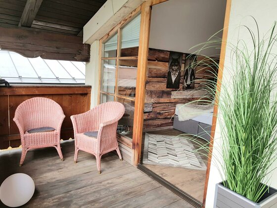 Balkonstühle Belle Kitz Zwei rosa Korbsessel auf einem Holzbalkon neben einer Glasfront zum Innenraum.
