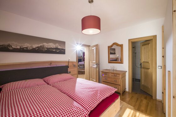 Schlafzimmer im Haus Skida Schlafzimmer mit Doppelbett, rot-weiß karierter Bettwäsche, Holzkommode und Blick ins Badezimmer.
