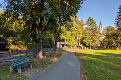 Parkweg in Steuerberg Asphaltierter Fußweg in einer Parkanlage mit Holzbänken, Bäumen und einer Kirche im Hintergrund.
