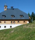 Dreistöckiges alpines Gebäude mit dunklem Metalldach und kleinem Glockenturm auf einem Grashang.