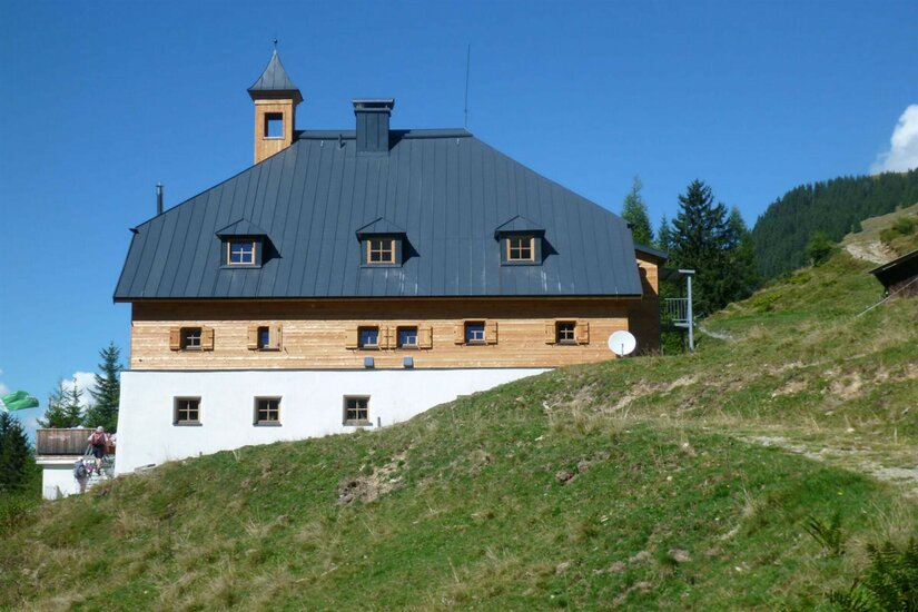 Dreistöckiges alpines Gebäude mit dunklem Metalldach und kleinem Glockenturm auf einem Grashang.