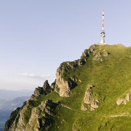 Ein hochgelegener Sendemast auf einem grünen Berghang, umgeben von felsigen Strukturen und einer weiten, offenen Landschaft.
