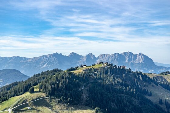 Hahnenkamm bei Aurach Blick auf bewaldete Bergrücken mit Gebäuden auf dem Gipfel vor einer schroffen Felskette unter blauem Himmel.