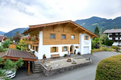 Zweistöckiges Landhaus mit Holzfassade im Obergeschoss und Bergen im Hintergrund.
