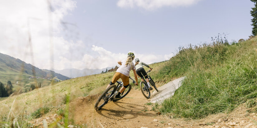 Zwei Mountainbiker fahren auf dem Sonnenrast Trail in Kitzbühel auf einem kurvenreichen Pfad durch eine grüne Landschaft unter blauem Himmel.