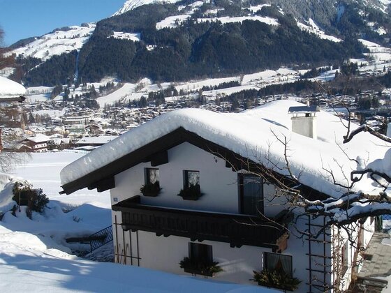 Haus Kolb im Winter Verschneites Haus mit Holzbalkon vor einer alpinen Bergkulisse und einer Ortschaft im Tal.