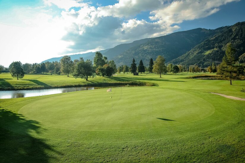 Gepflegtes Green auf einem Golfplatz mit Wasserhindernis vor bewaldeten Bergen unter bewölktem Himmel.