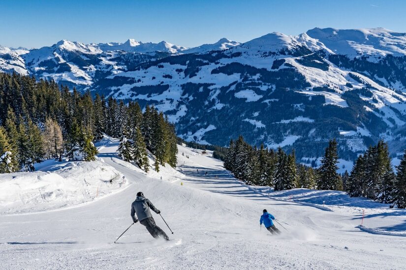 Zwei Skifahrer auf einer präparierten Piste vor verschneitem Bergpanorama und Nadelwald.
