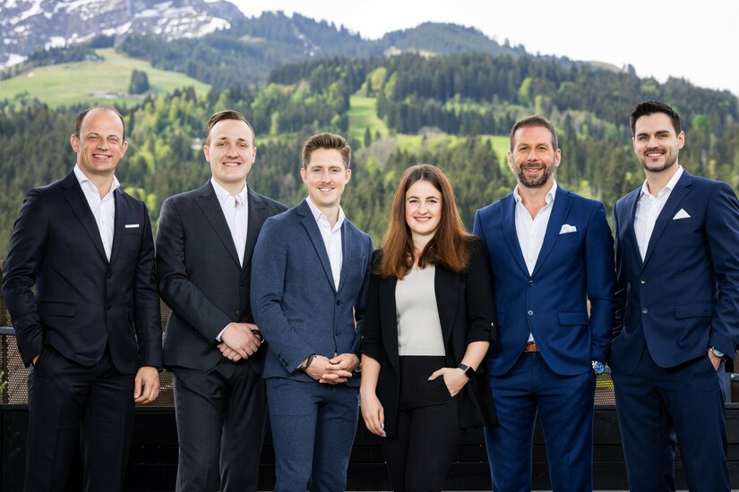 Team von Cum Laude Immobilia Sechs Personen in Business-Kleidung stehen nebeneinander vor einer Bergkulisse mit Wäldern.