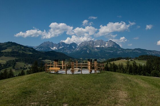 Aussichtspunkt am Reither Trail Eingezäunter Aussichtspunkt mit Holzbank auf einem Grashügel vor einem Gebirgspanorama unter blauem Himmel.