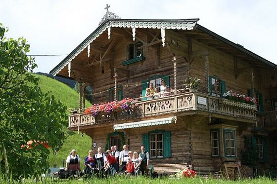 Traditionelles Holzhaus mit einer Gruppe von Menschen in Tracht davor und auf dem Balkon.