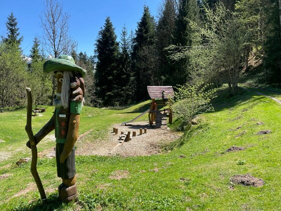 Holzfigur eines Wanderers auf einer Wiese vor einem Waldspielplatz mit Rutsche.