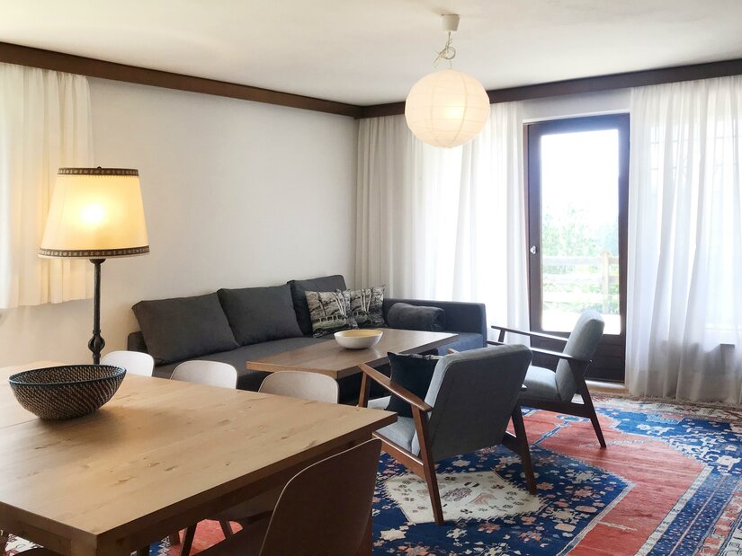 Wohnzimmer mit grauem Sofa, zwei Sesseln, Holztisch und buntem Teppich vor einer Balkontür.