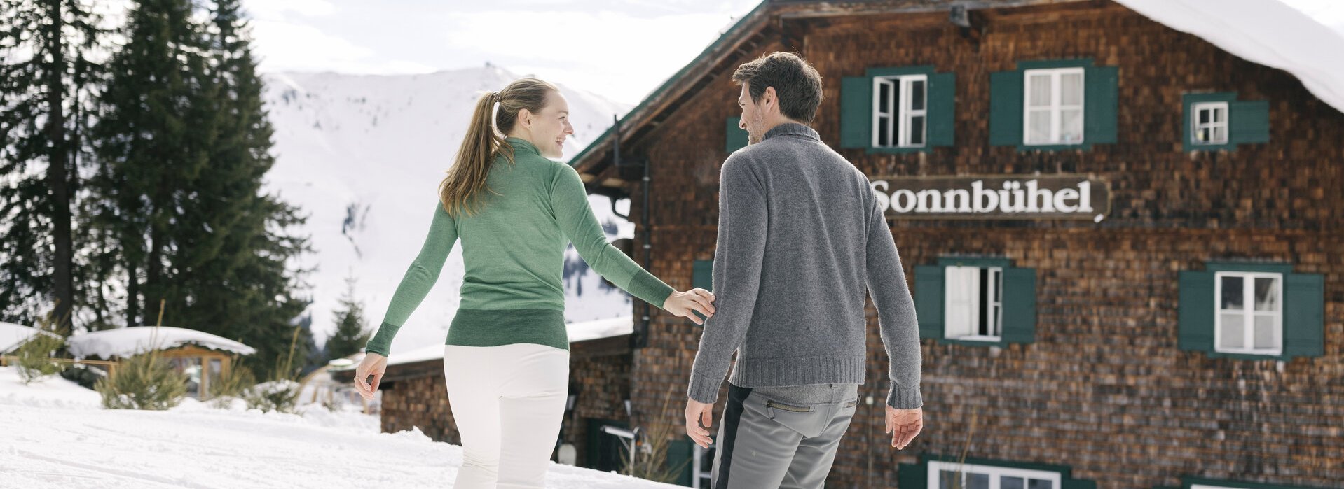 A couple in winter jumpers in front of the Sonnbühel mountain hut in the forest covered in white snow