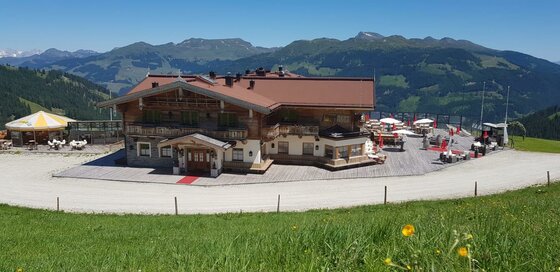 Sonnalm Alpinlodge Große Berghütte aus Holz und Stein mit Sonnenterrasse vor einem Gebirgspanorama.