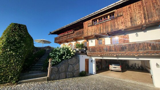 Chalet Nestwärme Außenansicht Alpines Holzhaus mit Balkon, Steintreppe und Garage unter blauem Himmel.
