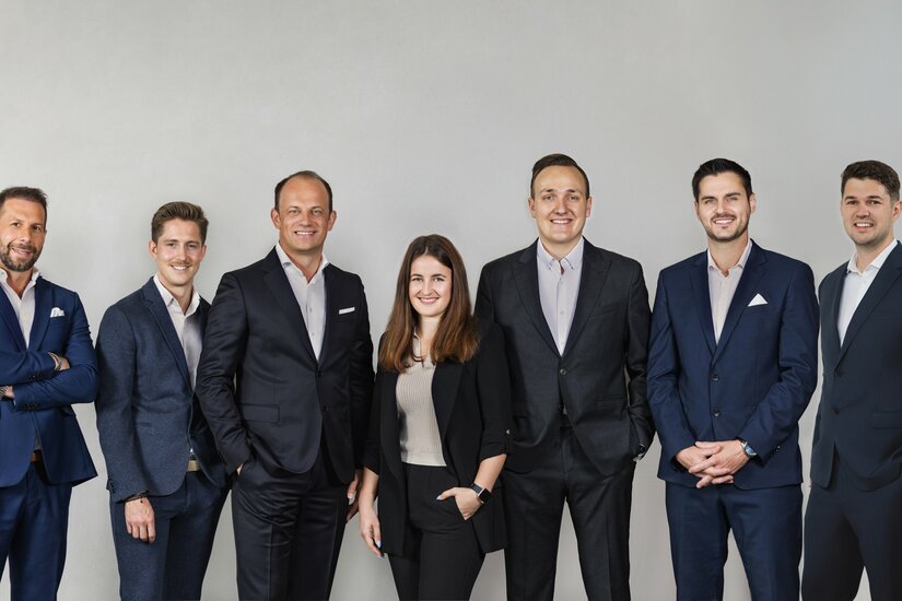 Sieben Personen in Business-Kleidung stehen nebeneinander vor einem grauen Hintergrund.