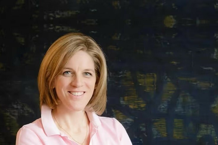 Dr. Petra Pölzelbauer Porträt einer lächelnden Frau mit blonden Haaren in einem rosa Poloshirt vor dunklem Hintergrund.