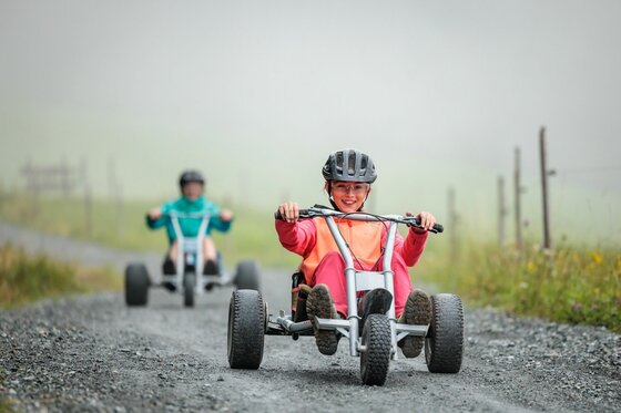 Zwei Personen fahren mit Mountaincarts auf einem Schotterweg in den Bergen.
