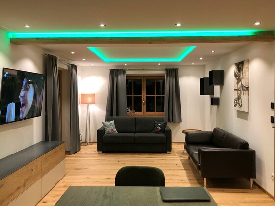 Modernes Wohnzimmer mit zwei dunklen Sofas, Fernseher und grüner LED-Deckenbeleuchtung.