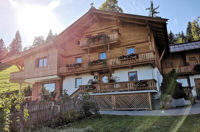 Mehrstöckiges Holzhaus im alpinen Stil mit Blumenbalkonen und weißem Erdgeschoss unter blauem Himmel.