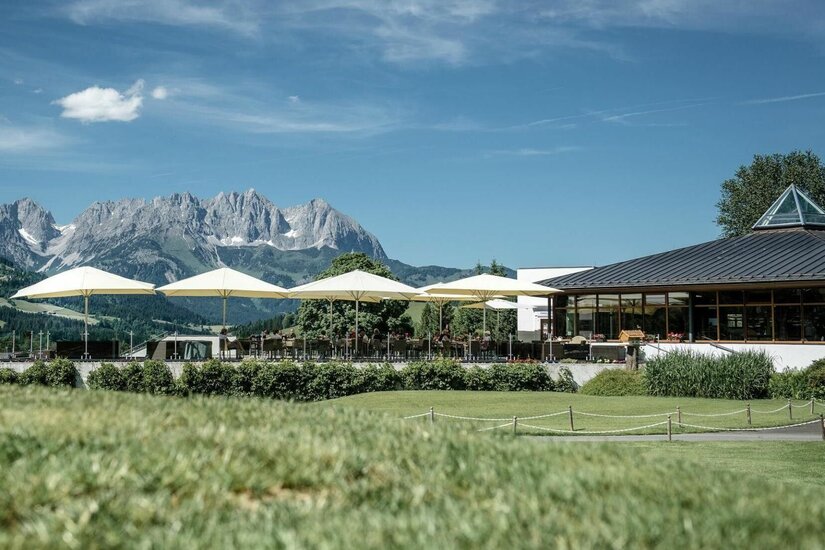 Golfrestaurant Schwarzsee Terrasse Sonnenterrasse eines Gebäudes mit weißen Schirmen vor einem Gebirgspanorama.