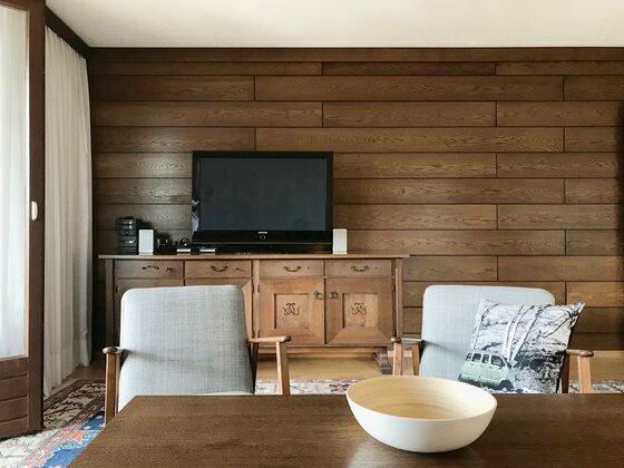 Wohnzimmer mit Holzvertäfelung, Fernseher auf Sideboard und zwei Sesseln vor einem Tisch.