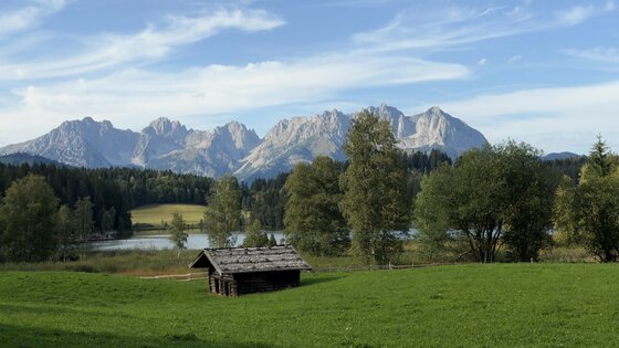 Holzhütte auf einer Wiese vor einem See und einem schroffen Gebirgsmassiv unter blauem Himmel.