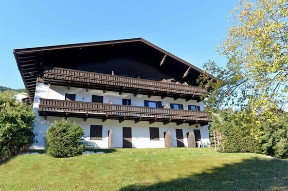 Appartementhaus Sommer Mehrstöckiges weißes Gebäude mit dunklen Holzbalkonen und flachem Satteldach vor blauem Himmel.
