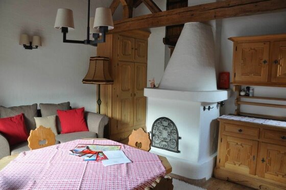 Wohnbereich Appartement 2 Rustikaler Wohnraum mit weißem Kachelofen, Holztisch und Sofa.