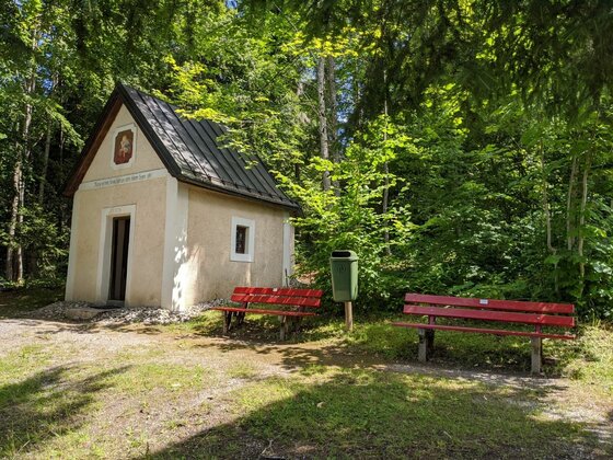 Waldkapelle am Sonnbergweg Kleine Kapelle mit Satteldach im Wald, davor zwei rote Holzbänke und ein Mülleimer.