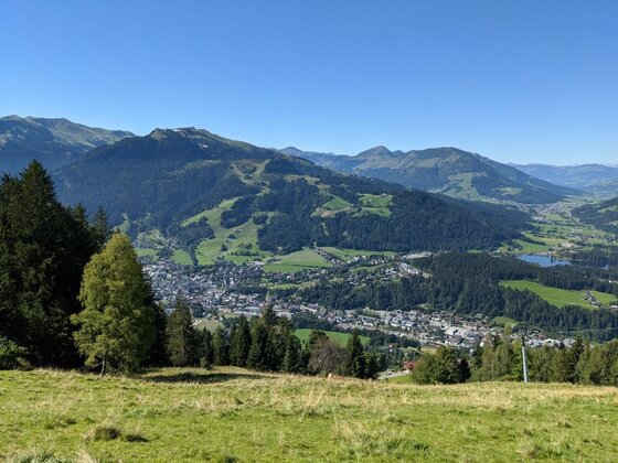 Blick ins Tal bei Kitzbühel Blick von einer Almwiese auf eine Stadt im Tal und bewaldete Berge unter blauem Himmel.