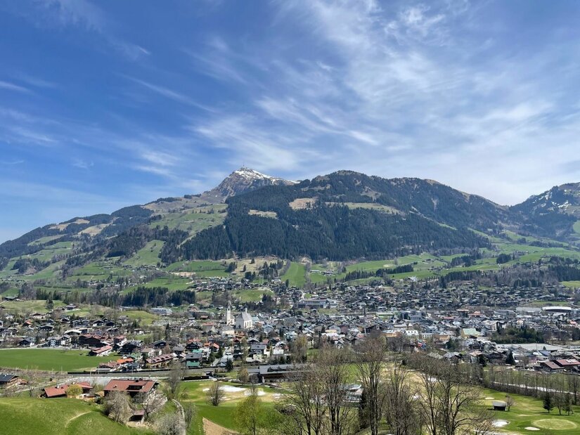 Blick auf eine Ortschaft im Tal vor bewaldeten Bergen unter blauem Himmel.