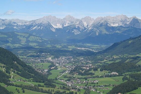 Blick von einem bewaldeten Hang auf ein Tal mit Siedlungen vor einer markanten, felsigen Gebirgskette.