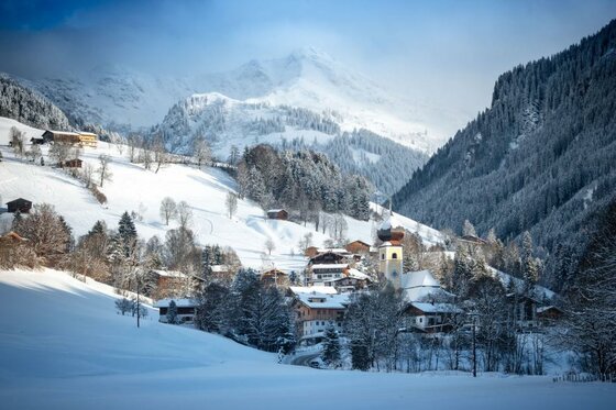 Verschneites Bergdorf mit Kirchturm vor bewaldeten Hängen und schneebedeckten Gipfeln.