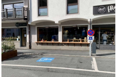 Behindertenparkplatz - Bichlstraße 22 Ein Laden mit großen Fenstern und einer Holzbank vor dem Eingang. Auf der Straße ist ein Parkplatz für Menschen mit Behinderungen markiert.