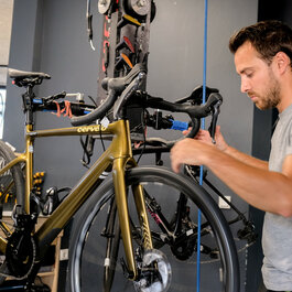 Ein Mann arbeitet konzentriert an einem goldenen Rennrad in einer Fahrradwerkstatt, umgeben von Werkzeugen und anderen Fahrrädern.