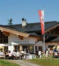 Außenansicht des Hahnenkamm Stüberls mit Gästen auf der Terrasse und Tiroler Fahne unter blauem Himmel.