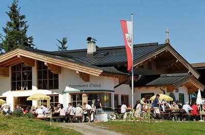 Hahnenkammstüberl im Sommer Außenansicht des Hahnenkamm Stüberls mit Gästen auf der Terrasse und Tiroler Fahne unter blauem Himmel.