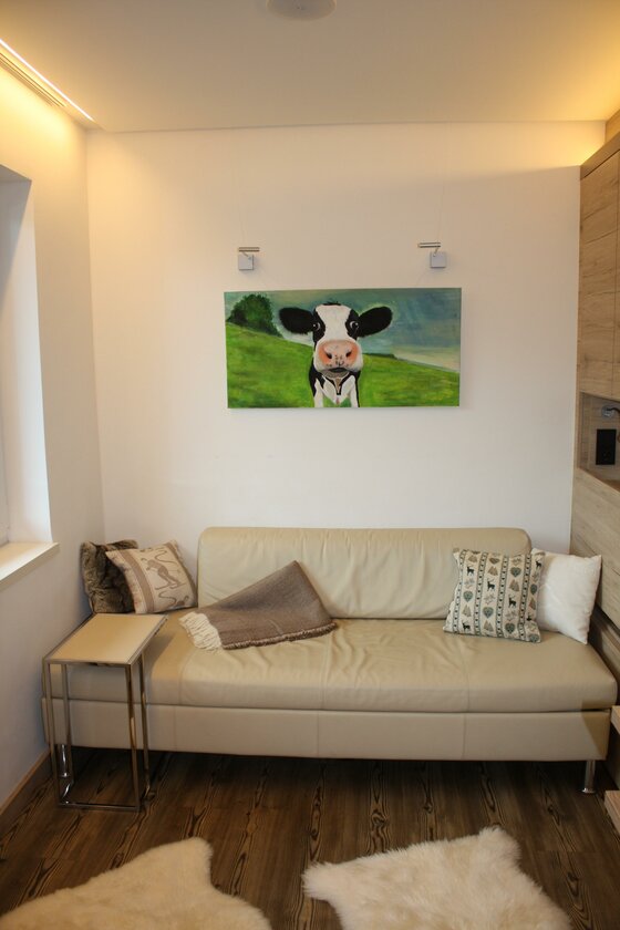 Modernes Wohnzimmer mit hellem Ledersofa, Kuh-Gemälde an der Wand und dunklem Holzboden mit Fellteppichen.