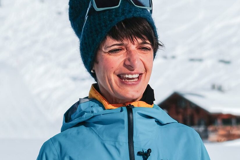 Eine lächelnde Person trägt eine warme Jacke und eine Mütze in den Bergen. Im Hintergrund sind schneebedeckte Hänge zu sehen.